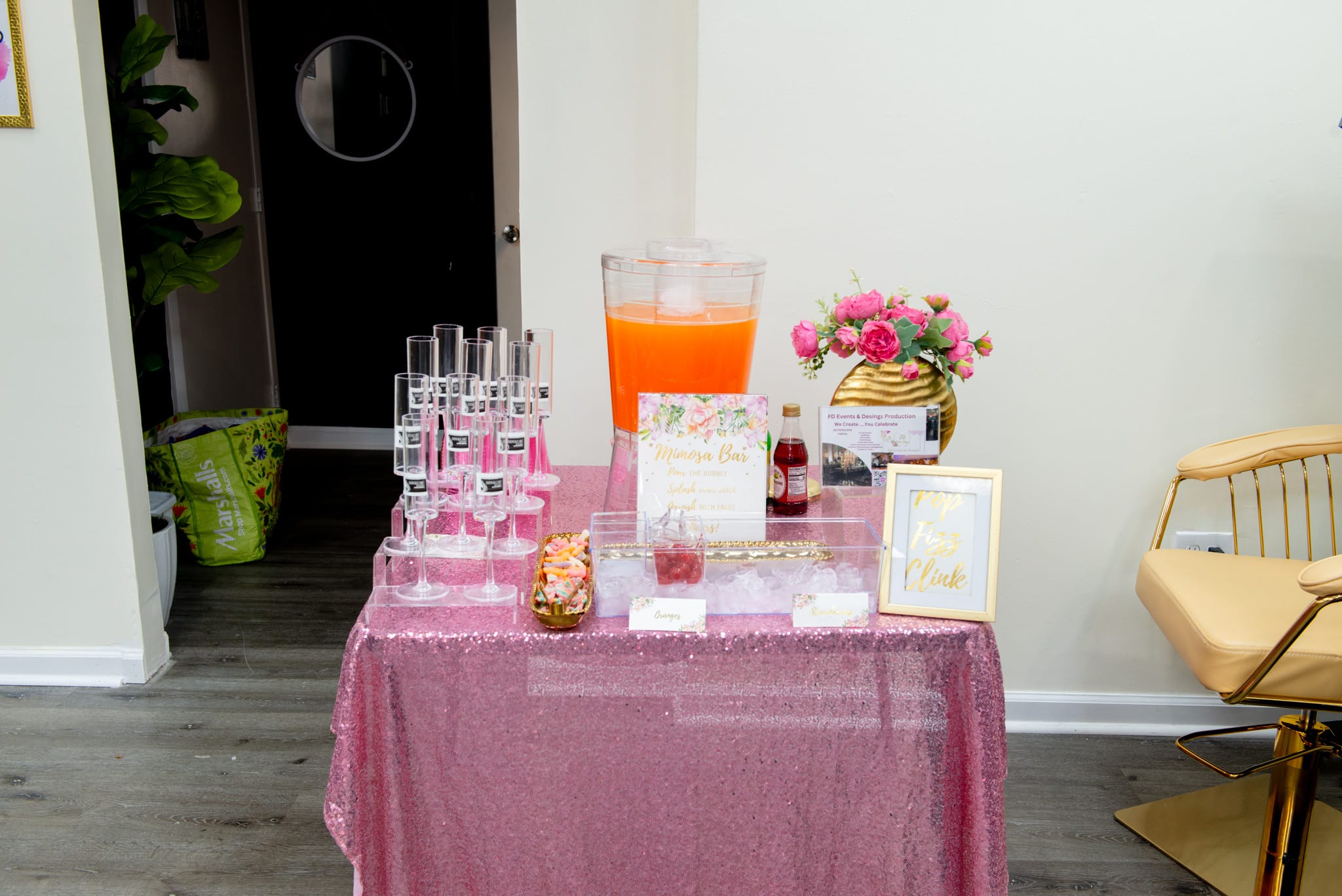 Mimosa bar station at spa party — pink sparkle tablecloth, champagne flutes, fresh juice dispenser, flower arrangement